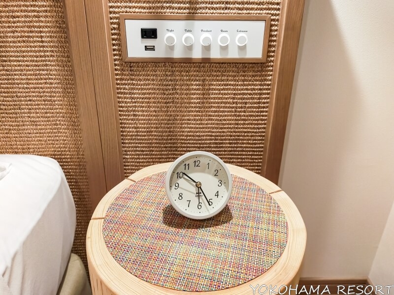 木の丸太でできているベッドサイドテーブルの上に丸いアナログ時計、ベッドヘッドにダイヤル式のスイッチ類